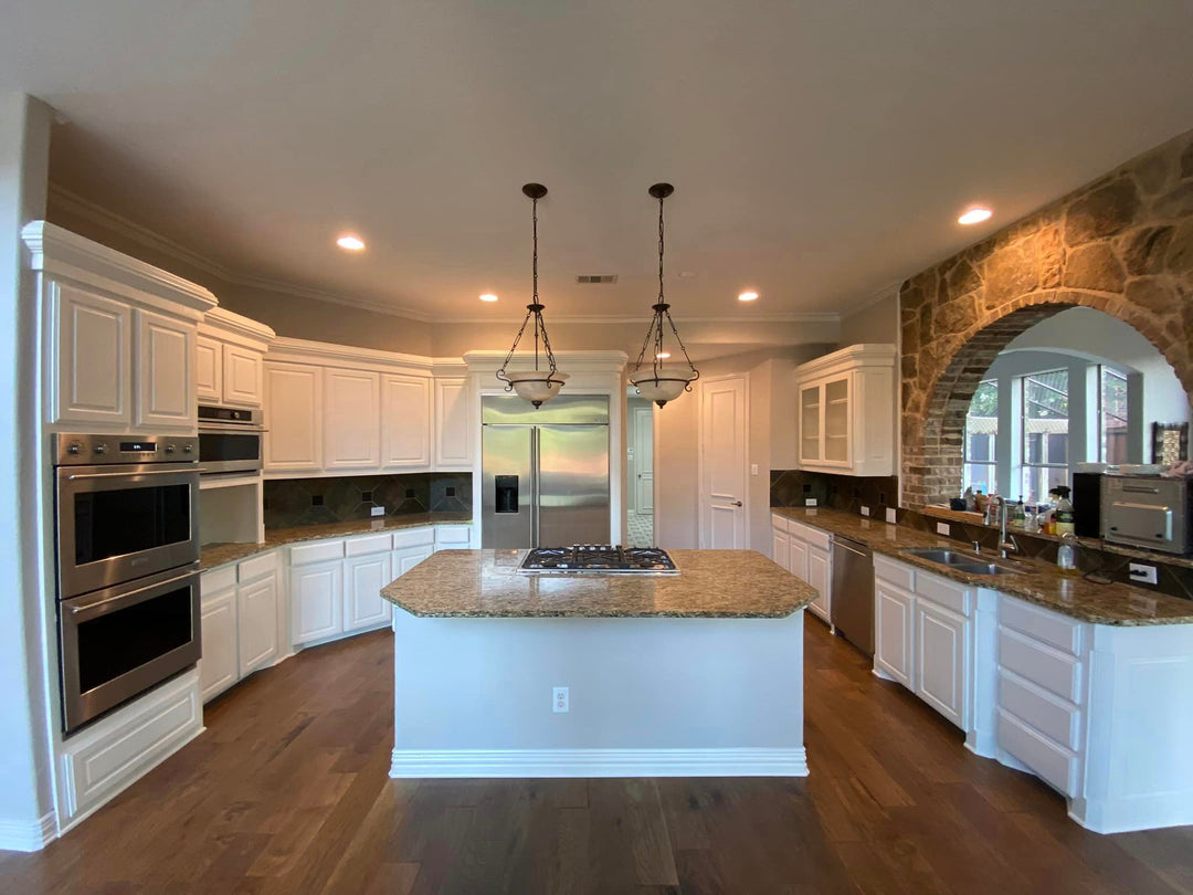 photo of a newly finished kitchen with white cabinets wood floors stainless steel appliances, an island in the middle and tan marble countertops